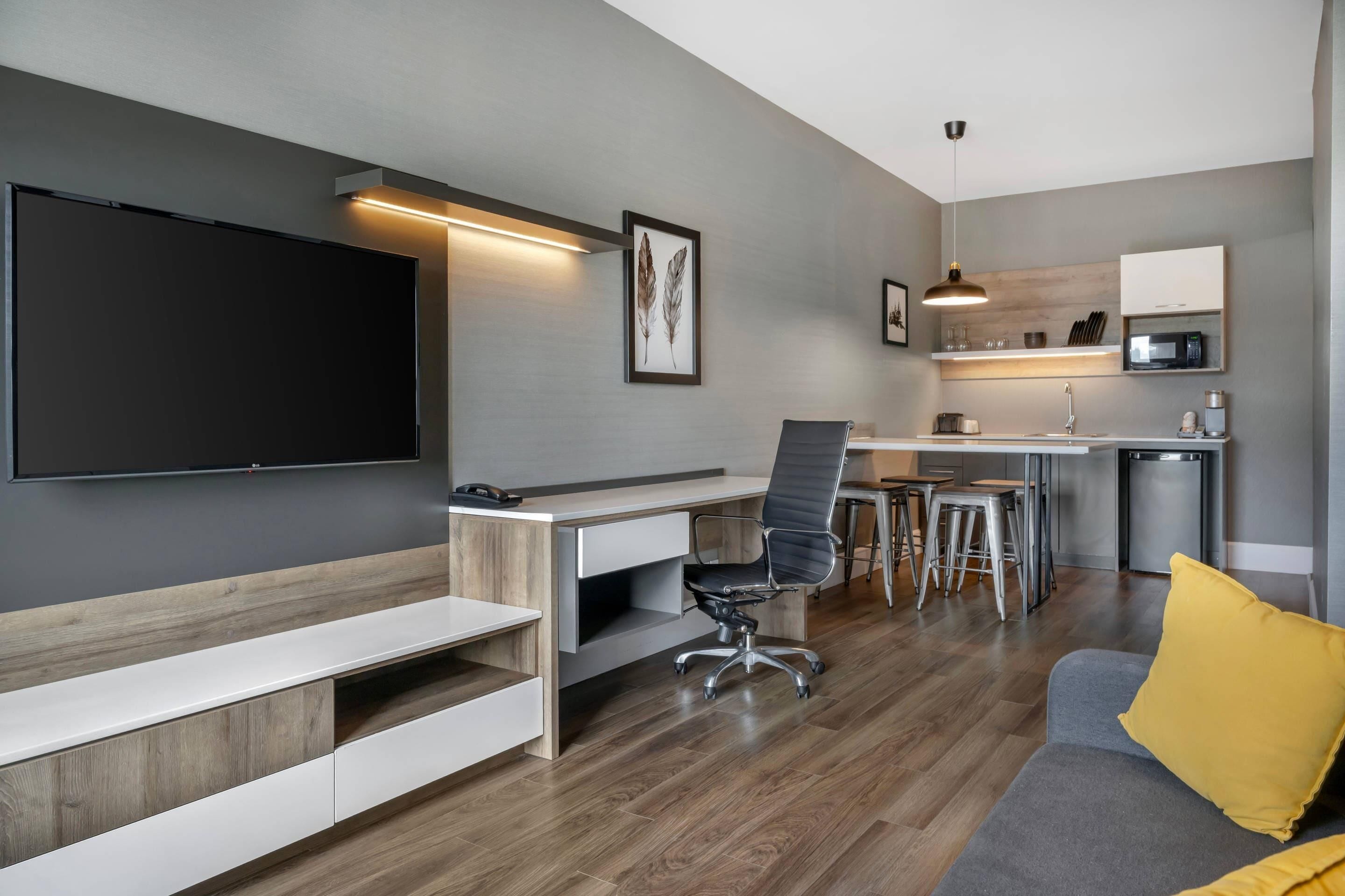 A modern living space with a flat-screen TV, a desk, a chair, a sofa, and a kitchen area with a microwave.