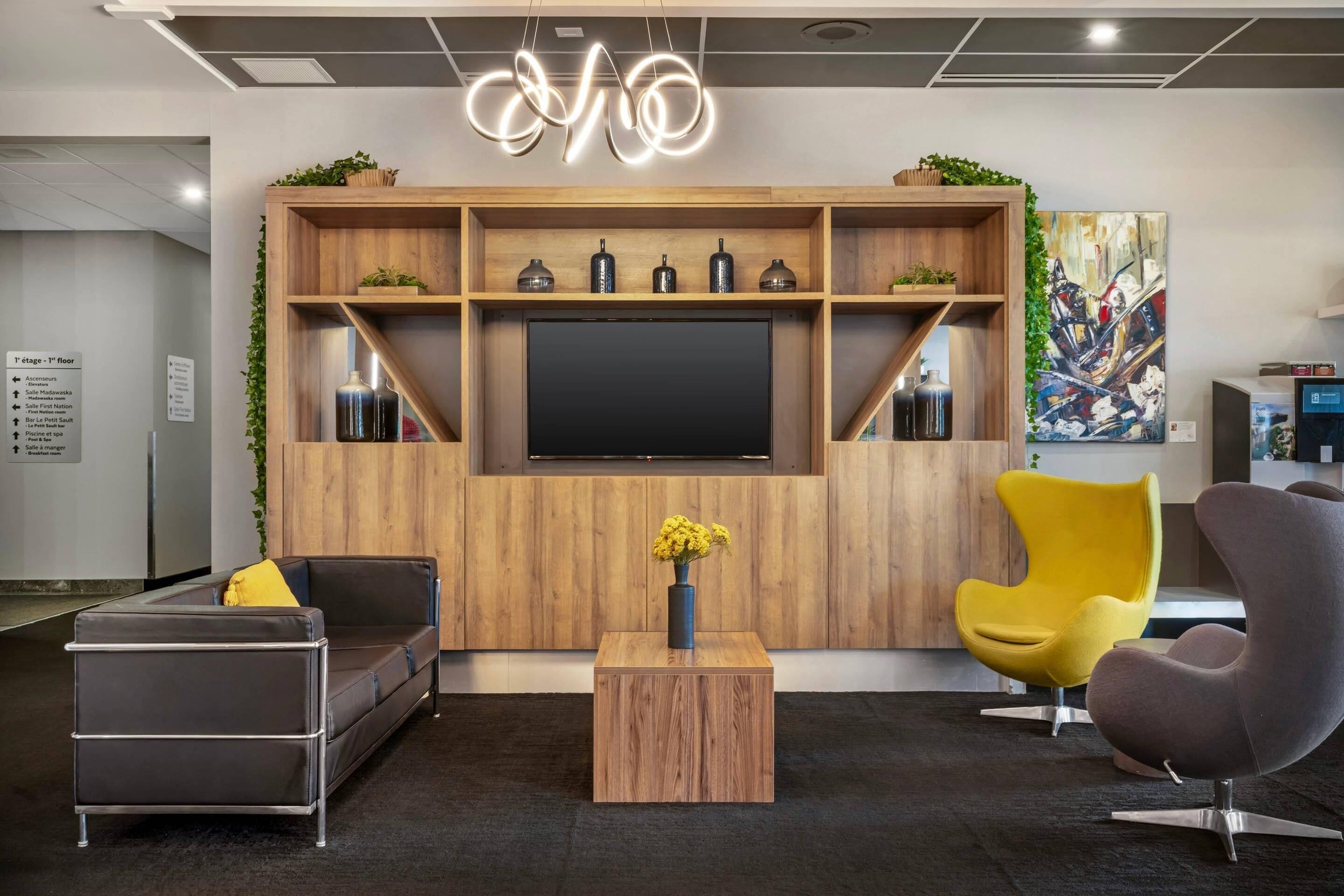 Un espace de détente moderne pour les bureaux, doté d’un grand téléviseur à écran plat, d’étagères en bois ornées d’objets décoratifs et de sièges contemporains.