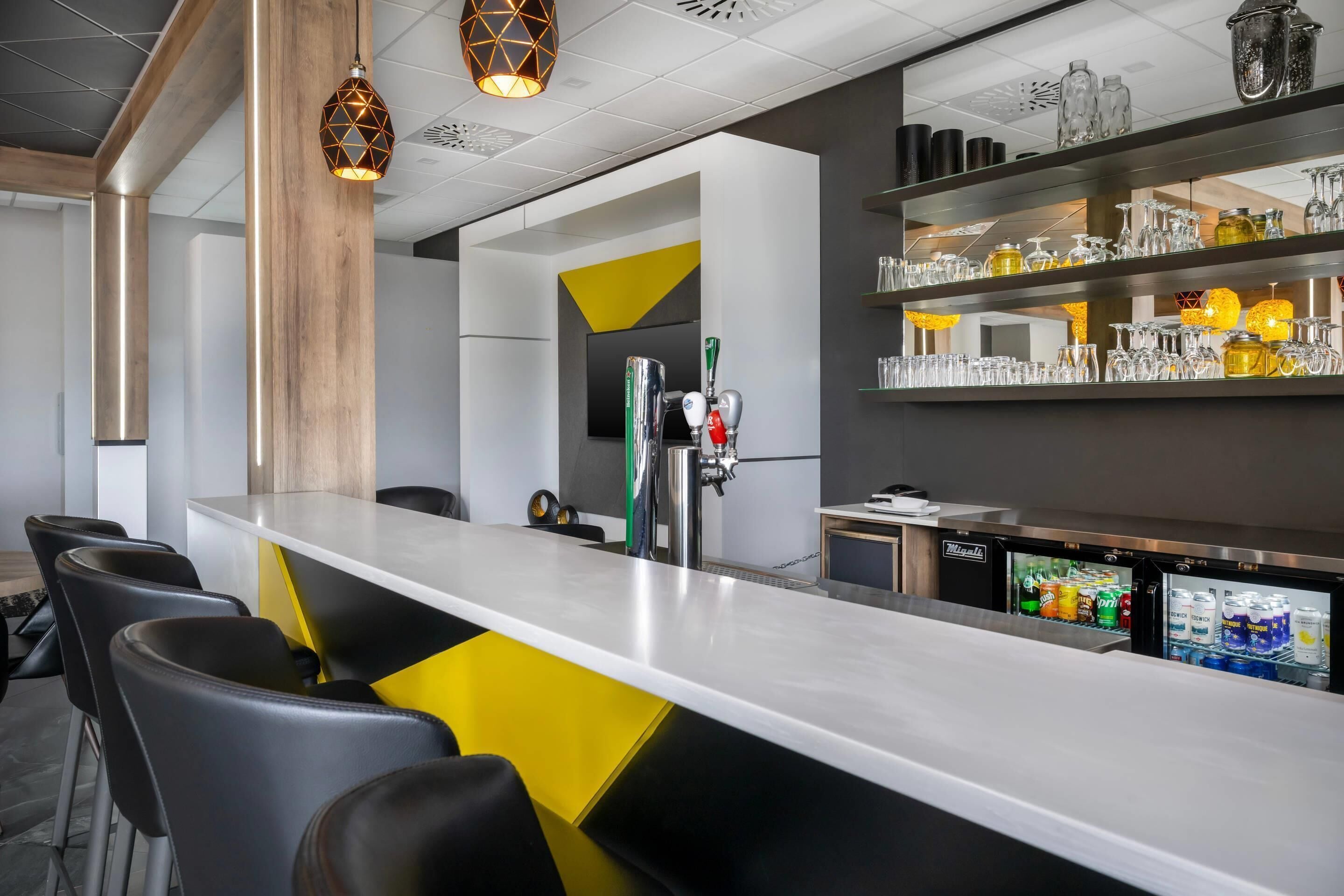 A modern bar area with a long counter, bar stools, a refrigerator stocked with beverages, and a selection of glassware on shelves.