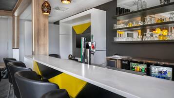 A modern bar area with a long counter, bar stools, a refrigerator stocked with beverages, and a selection of glassware on shelves.