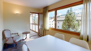 A hotel room with a large window offering a mountain view, a dining table with chairs, and a small round table with a chair. - The spacious 3 room apartment is located in a multi - family house at the villag (Celerina/Schlarigna)