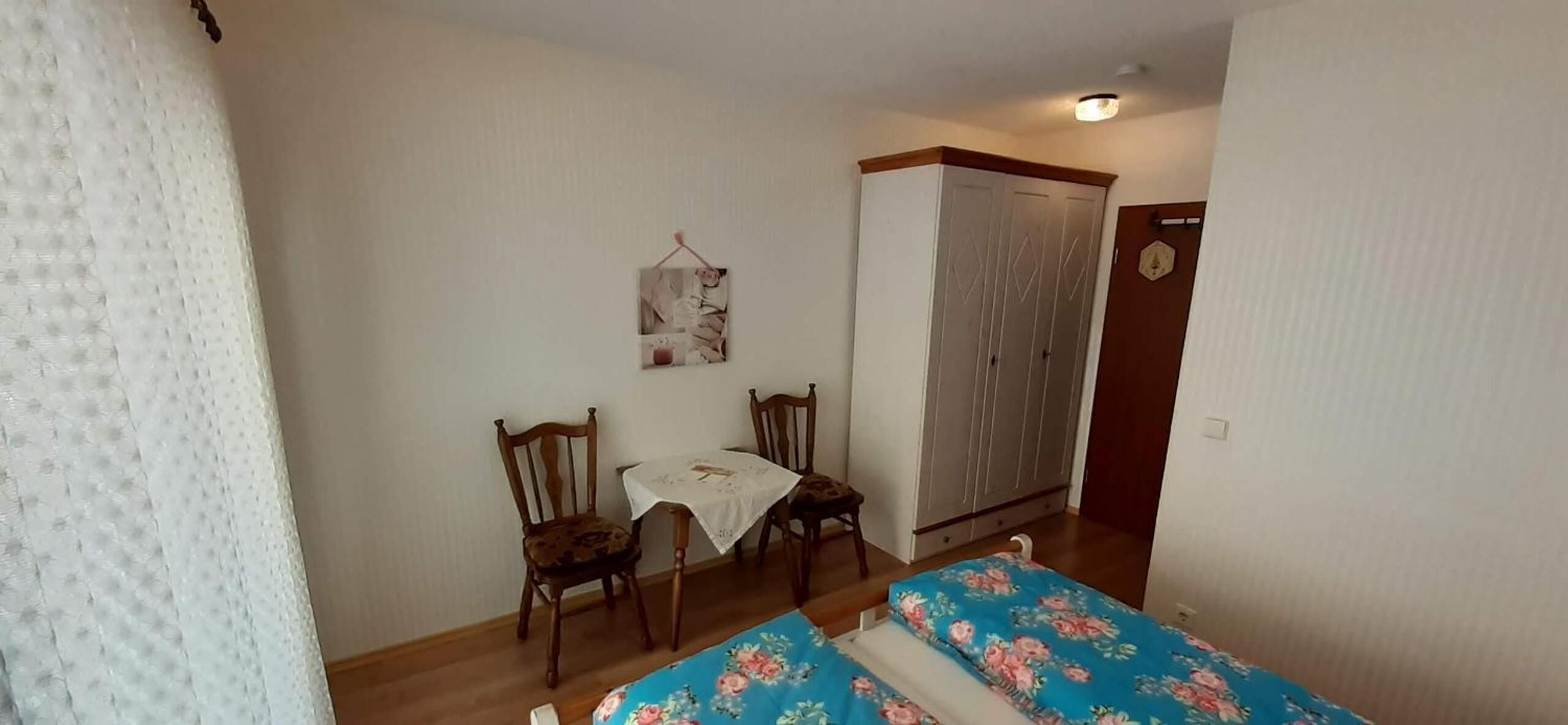 A small bedroom with a bed, two chairs, a table, and a wardrobe.