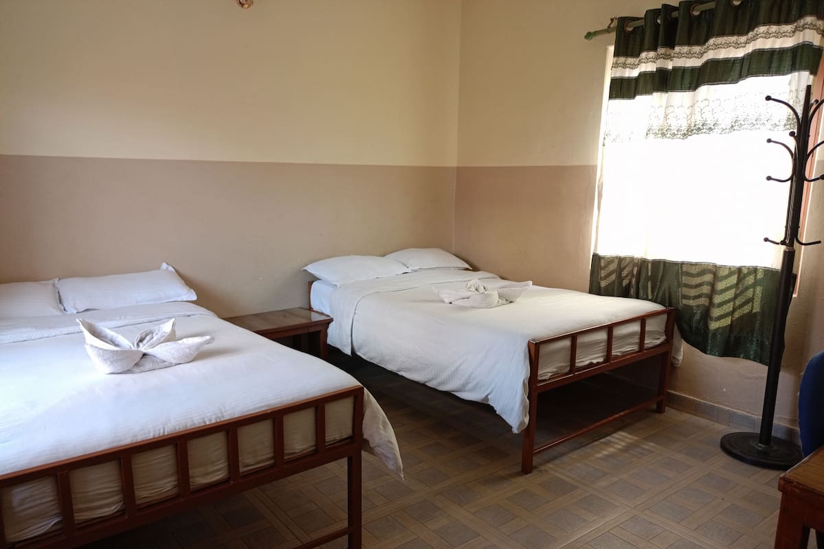 A hotel room with two single beds, a wooden floor, a window with a green curtain, and a coat rack.