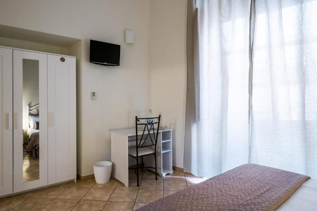 A bedroom with a bed, a desk, a chair, a wardrobe, and a TV on the wall.