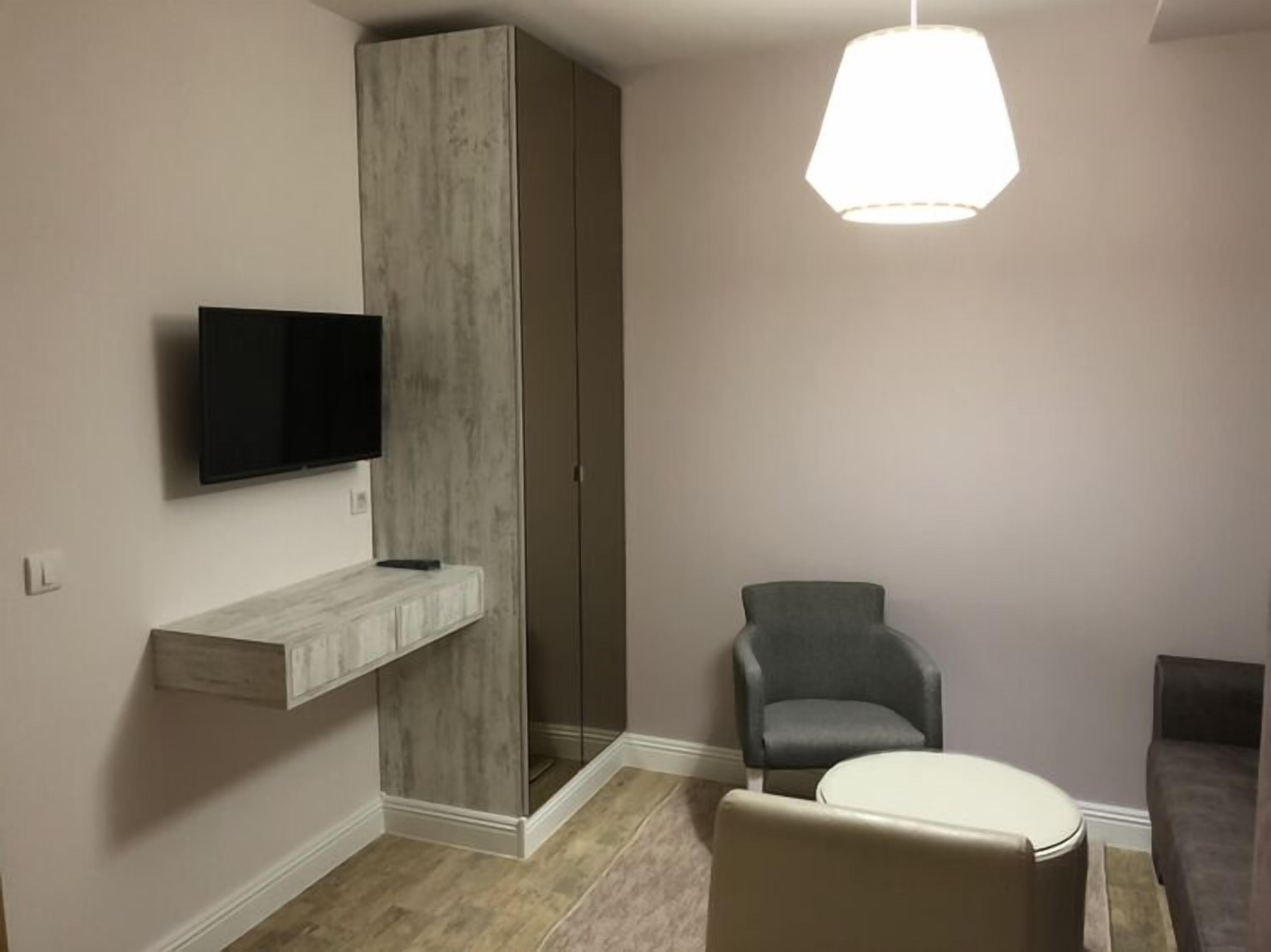 Una sala de estar moderna con un sofá gris, un sillón gris, un reposapiés blanco, un televisor montado en un estante de madera y una lámpara colgante.