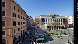 Un hotel con un patio central, un edificio grande con un banderín azul y una calle con coches aparcados y peatones.