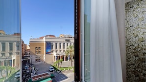 Vista desde una ventana con cortina floral, que da a una calle con edificios, vehículos estacionados y un camión verde.