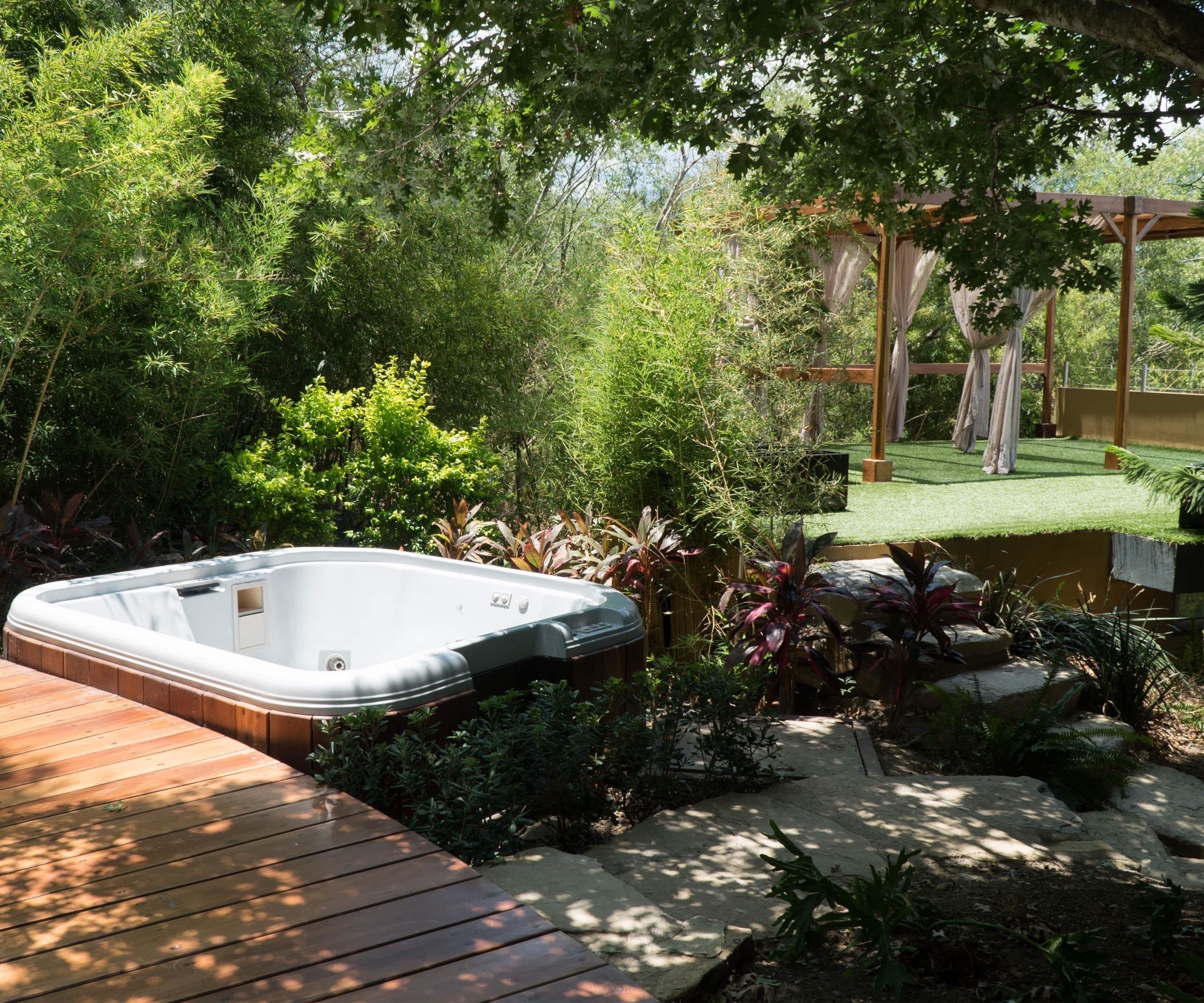 Un jacuzzi en un jardín exuberante con una terraza de madera, rodeado de diversas plantas y una estructura de pérgola.