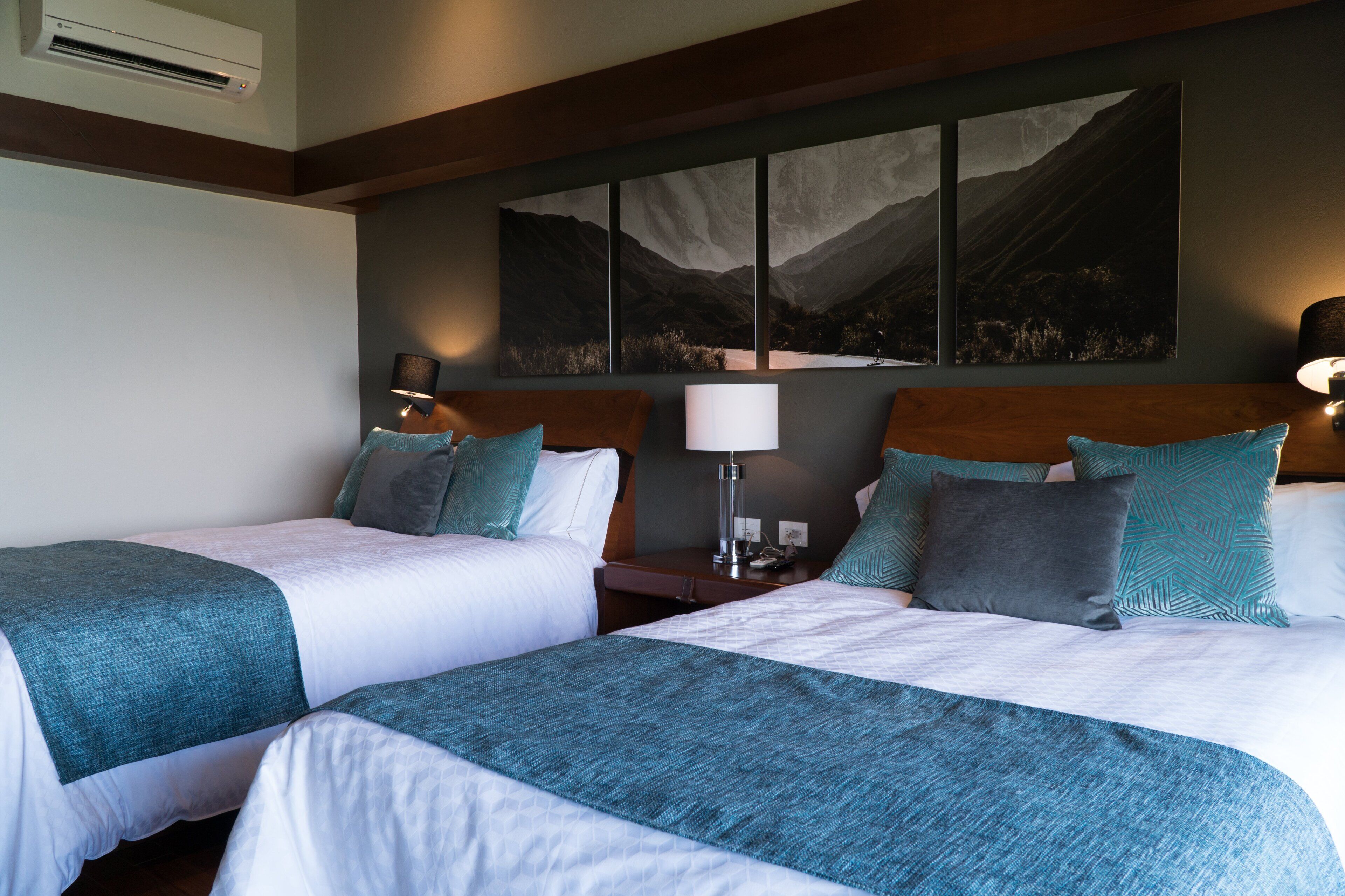 A hotel room with two beds, a large mountain view painting, and a nightstand with a lamp.