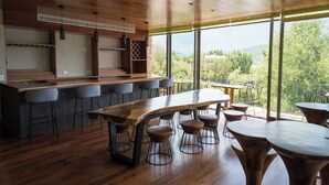 A modern bar area with a long wooden table and multiple stools, large windows offering a view of greenery and mountains. - Gamma Monterrey Rincón de Santiago. (Santiago)