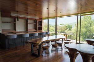 Una zona de bar moderna con una mesa de madera larga y varios taburetes, amplias ventanas que ofrecen vistas a zonas verdes y montañas.