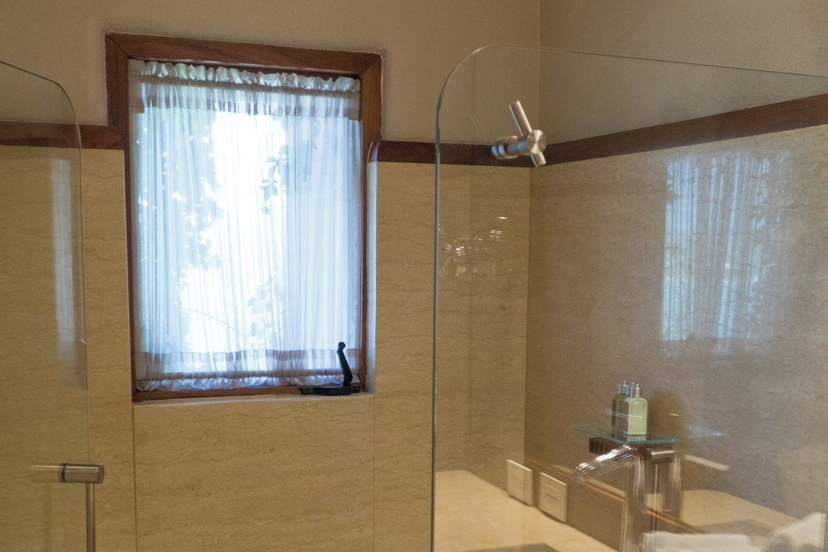 A modern bathroom with a glass-enclosed shower, a vanity with a mirror, and a window with sheer curtains.