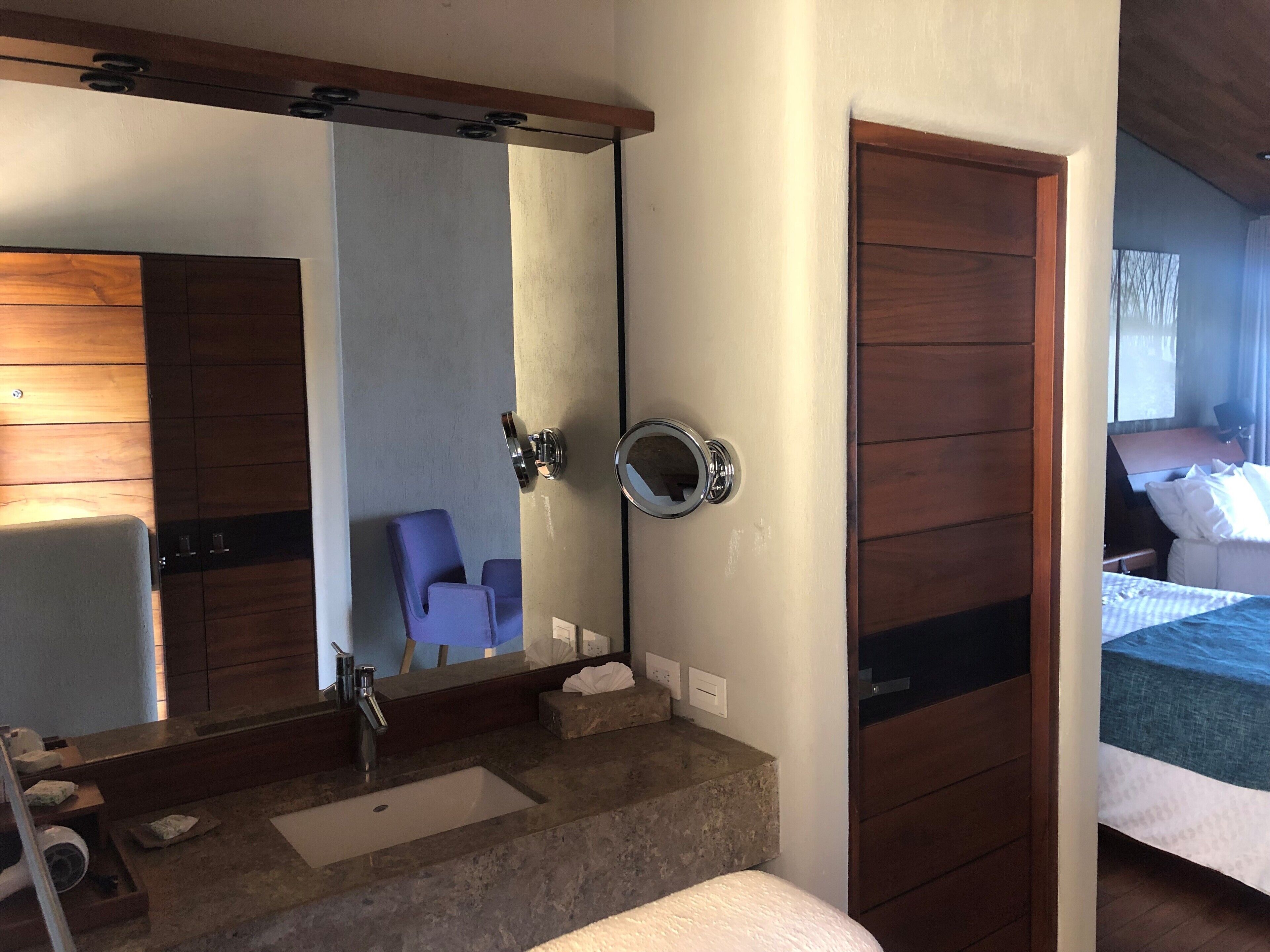 A hotel room with a large mirror, a wooden wardrobe, a bathroom with a sink and a towel rack, and a bed with a blue bedspread.