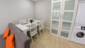Un espace de vie compact comprenant une table à manger, des chaises, un canapé avec des coussins orange, un meuble blanc avec des portes en verre et un lave-linge.