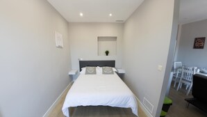 A modern bedroom with a large bed, bedside tables, a small plant, and a view into a dining area with chairs and a table.