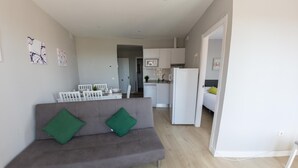 Una sala de estar moderna con un sofá gris y cojines verdes, una zona de cocina con electrodomésticos blancos y una mesa de comedor con sillas.