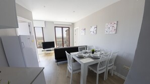 Una cocina moderna con armarios blancos, un comedor con una mesa blanca dispuesta para cuatro personas, y un sofá negro al fondo.