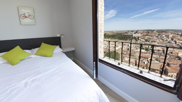 Un dormitorio con cama, almohadas verdes, cabecero, mesita de noche, un cuadro enmarco y un balcón con vista a la ciudad.