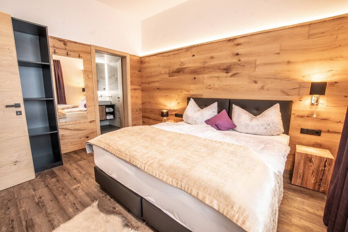 A modern bedroom with a wooden headboard, a large bed with a beige blanket, two bedside lamps, and a built-in wardrobe with a mirror.