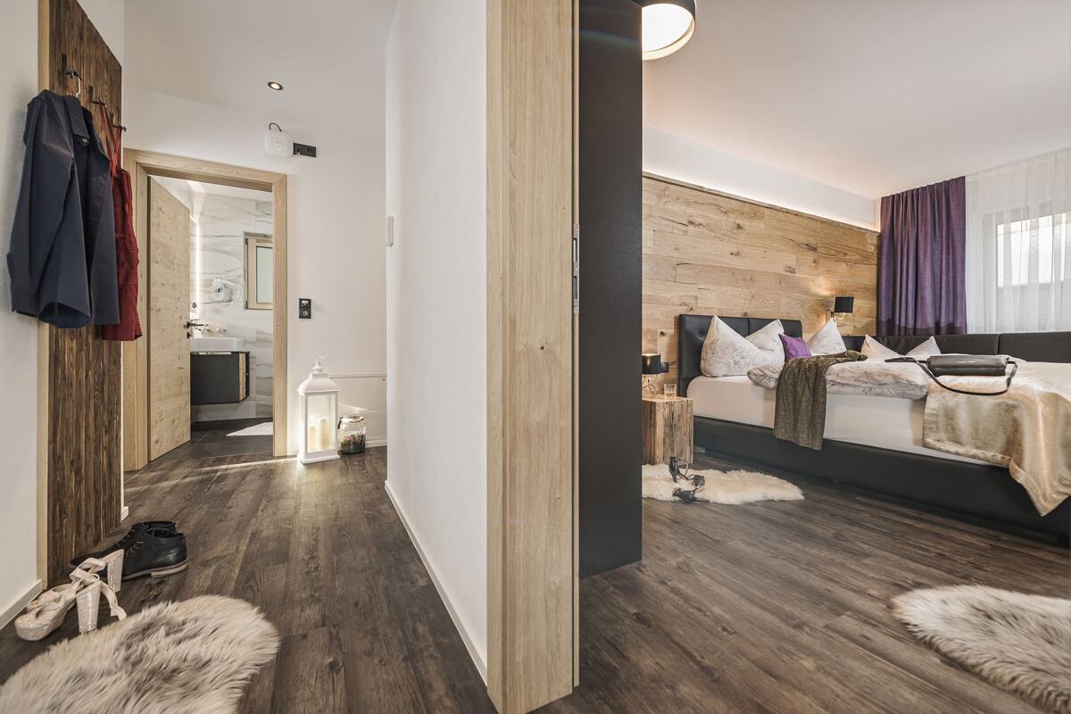 A modern hotel room with a wooden wall, a bed with multiple pillows, a nightstand, a lamp, and a view of the bathroom.