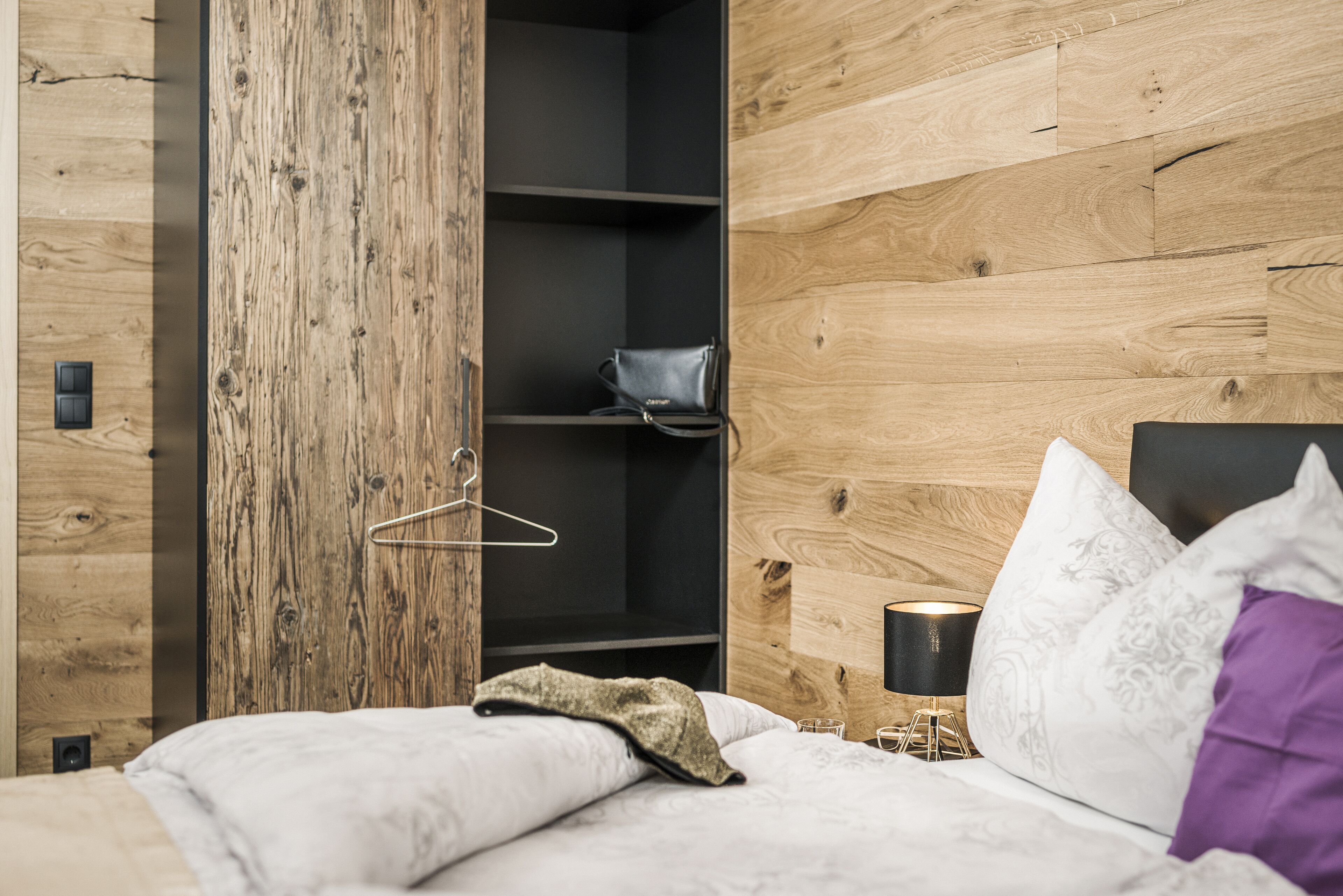 Un dormitorio con pared de madera, una cama con ropa de cama blanca, un bolso colgante y una mesita de noche con lámpara.