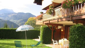 Un patio con due poltrone verdi e un tavolo, un balcone in legno con fioriere e una vista sulle montagne sullo sfondo.