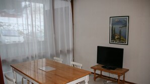 A dining area with a wooden table and chairs, a television on a stand, and a window with sheer curtains. - Studio Le Colombier N ° 18 (Aillon-le-Jeune)