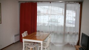 A room with a wooden dining table, white chairs, a TV, and a view through sheer curtains. - Studio Le Colombier N ° 18 (Aillon-le-Jeune)