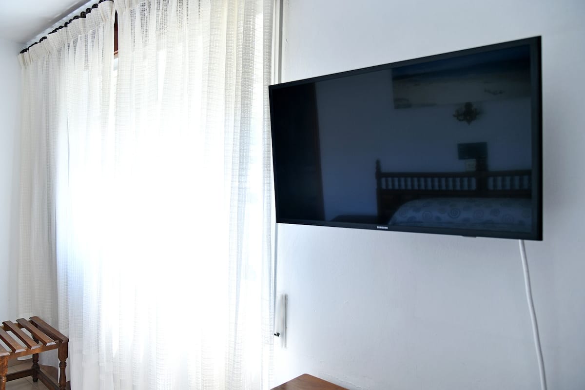Une pièce avec un téléviseur fixé au mur, une chaise en bois et une fenêtre ornée de rideaux légers.