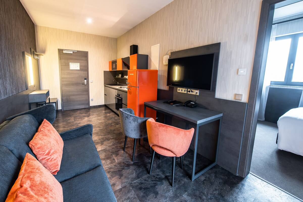 Ein modernes Hotelzimmer mit einem dunkelgrauen Sofa, zwei Sessel, einer kleinen Küchenzeile und einem an der Wand befestigten Fernseher.