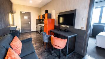Ein modernes Hotelzimmer mit einem dunkelgrauen Sofa, zwei Sesseln, einer kleinen Küchenzeile und einem an der Wand befestigten Fernseher.