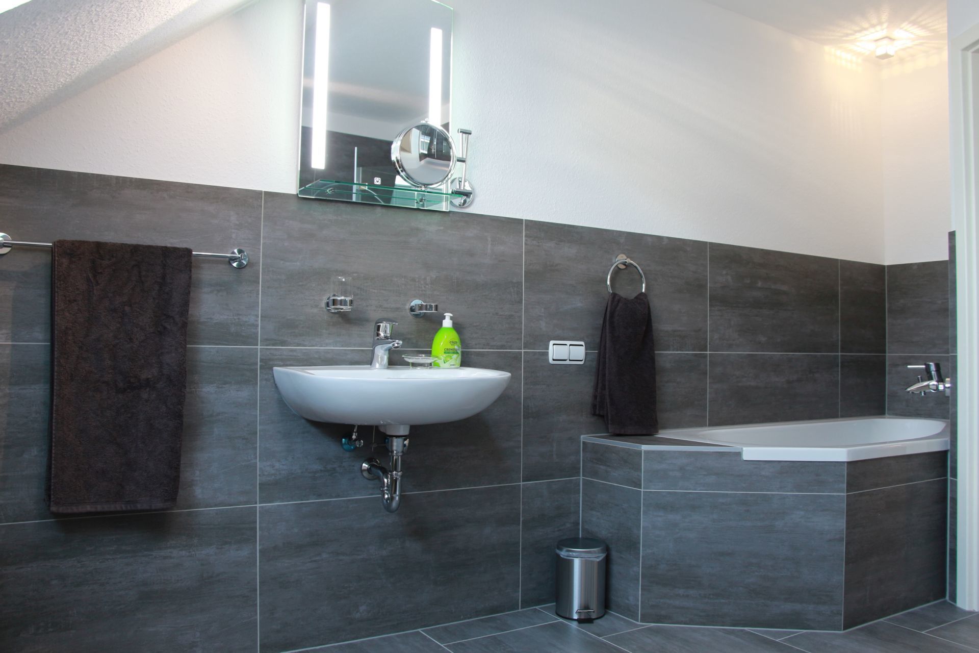A modern bathroom with a white sink, a large mirror, a glass-enclosed shower, and dark grey tiles.