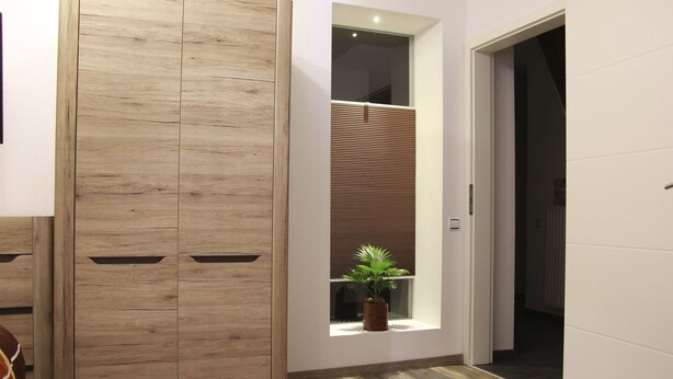Une pièce avec une armoire en bois, une fenêtre avec des stores, une plante en pot et une télécommande posée sur le sol.