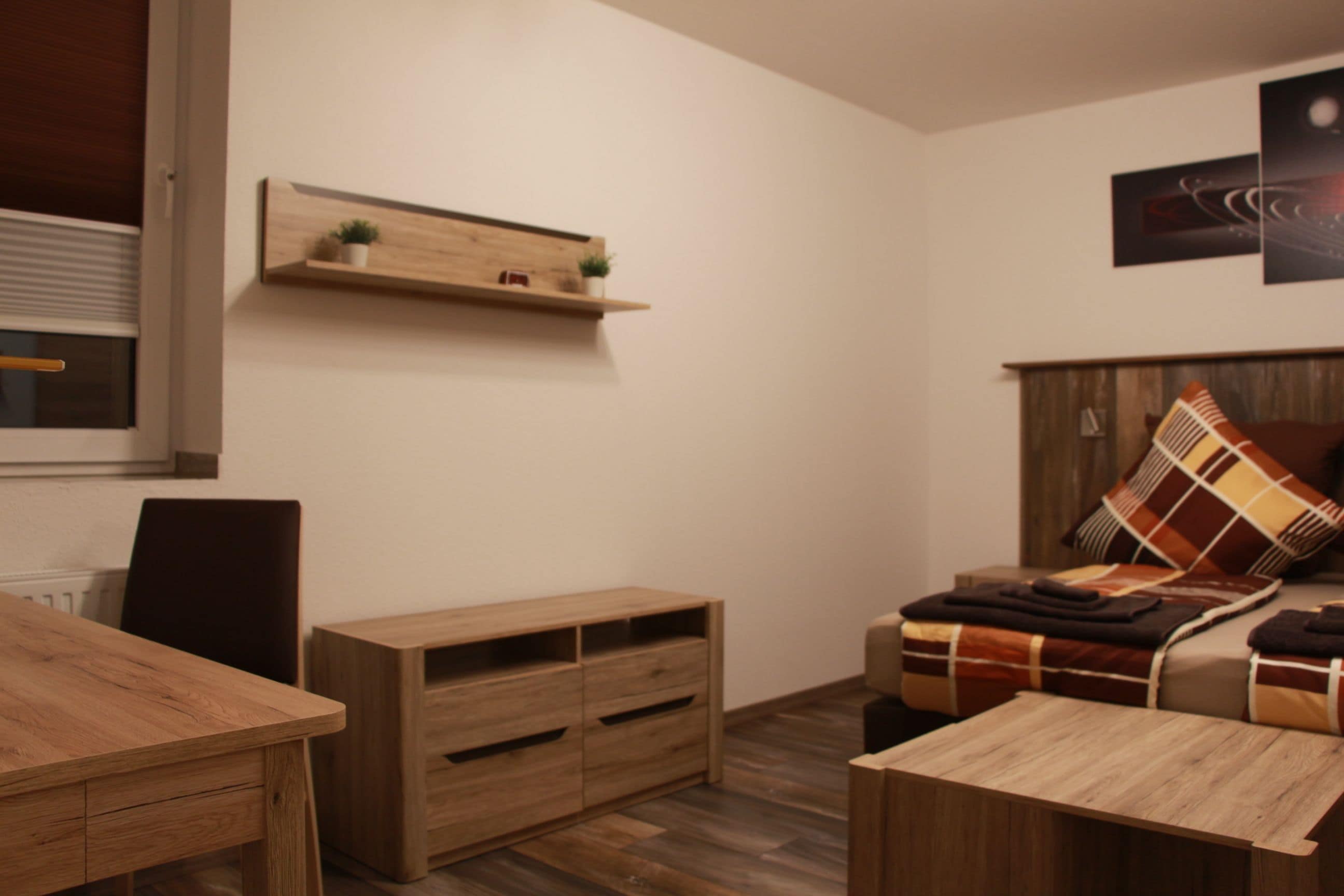 A modern living room with a wooden dining table, a sofa with a plaid blanket, a wooden cabinet, and a wall-mounted shelf with a plant.