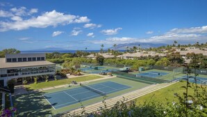 A tennis court complex with multiple courts, a building with large windows, and a view of the ocean and mountains in the background. - Traveler Favorite! Spacious, comfortable, updated 5-STAR Wailea Retreat. 🏝  (Wailea)