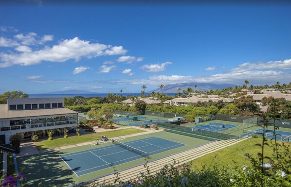 A tennis court complex with multiple courts, a building with large windows, and a view of the ocean and mountains in the background.