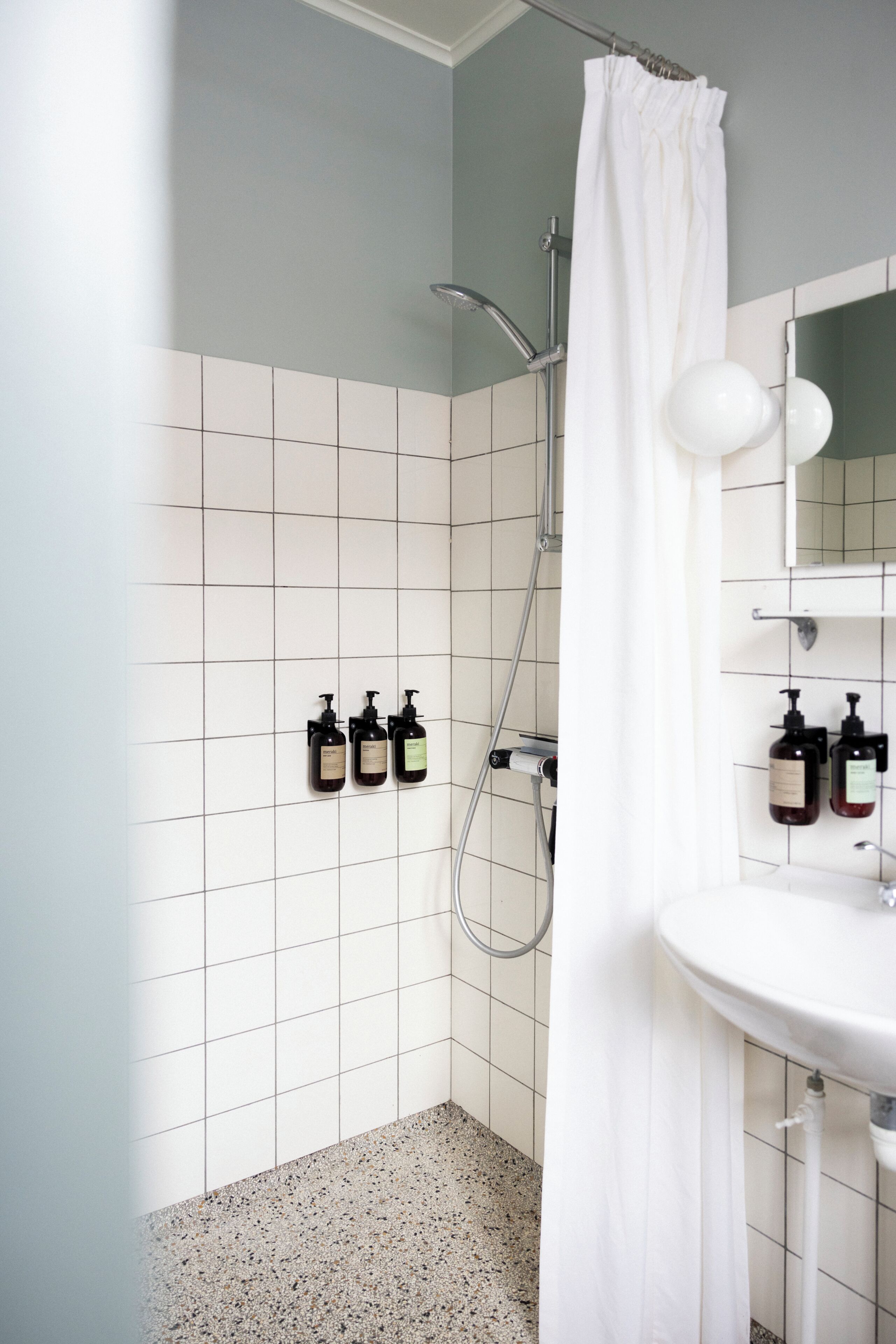 Un baño con azulejos blancos, ducha, lavabo y dos botellas de champú en la pared.