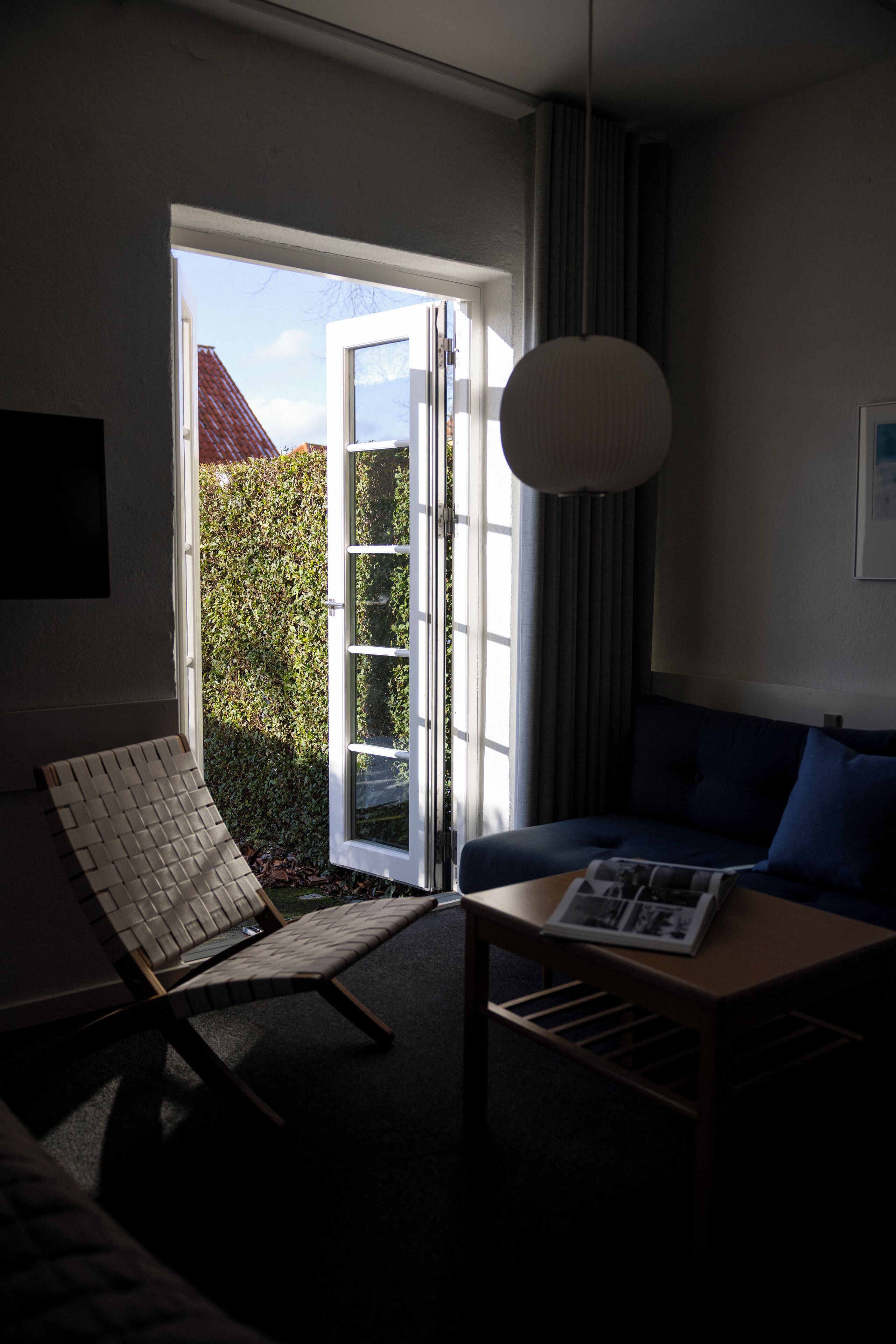 A room with a sofa, a table with a magazine, a chair, and a large window with a view of a hedge and a building.
