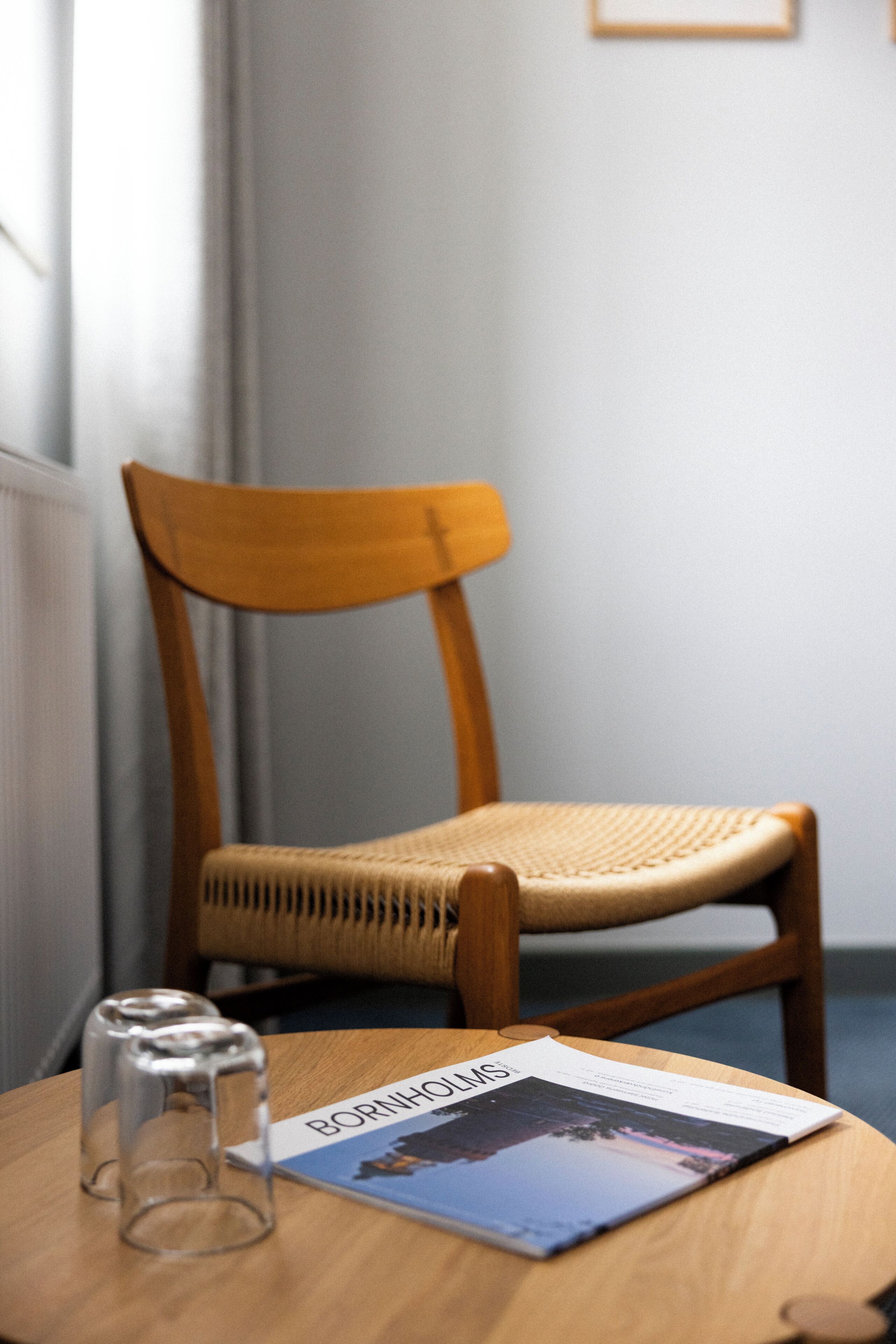 A wooden chair with a woven seat, a table with a magazine and glasses, and a window with curtains.