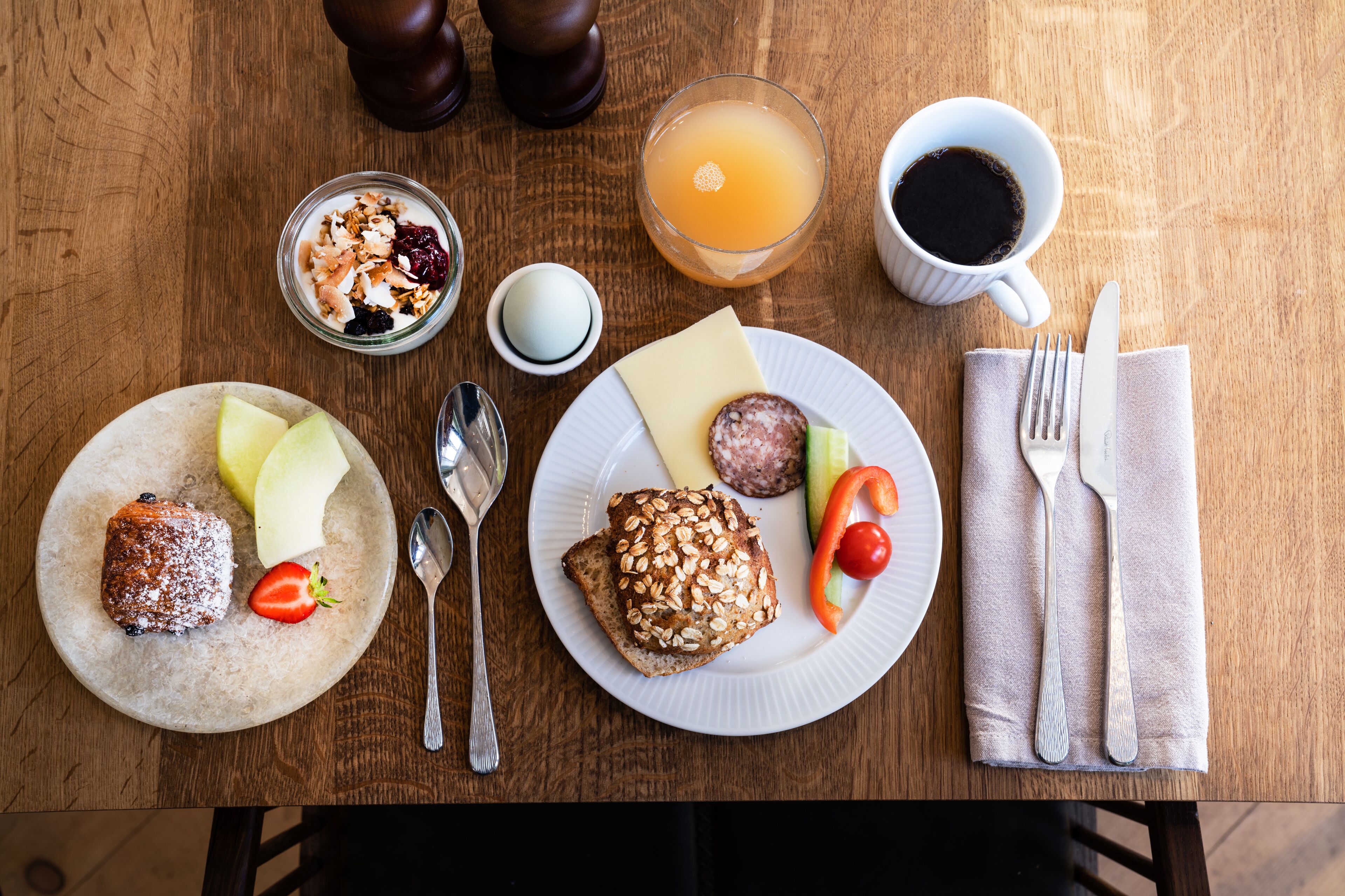 Une table de petit-déjeuner avec divers aliments, un verre de jus d’orange, une tasse de café et des couverts.