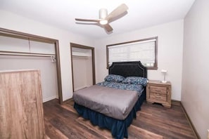 Une chambre à coucher comprenant un lit, une table de chevet, un ventilateur de plafond, une fenêtre avec des stores et une armoire.