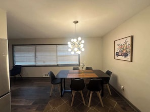A dining area with a table, chairs, a chandelier, a window with blinds, and a wall-mounted picture.