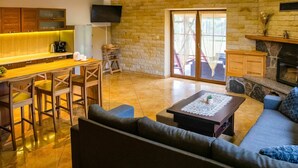 A modern living room with a sofa, coffee table, and a fireplace. There is a kitchen area with a bar and stools. The room has a stone wall and a large sliding glass door leading to an outdoor area.
