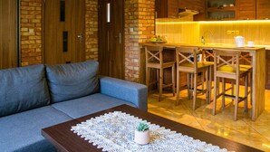 A modern living room with a grey sofa, a wooden coffee table with a lace table runner, and a kitchen area with wooden cabinets and a bar stool.