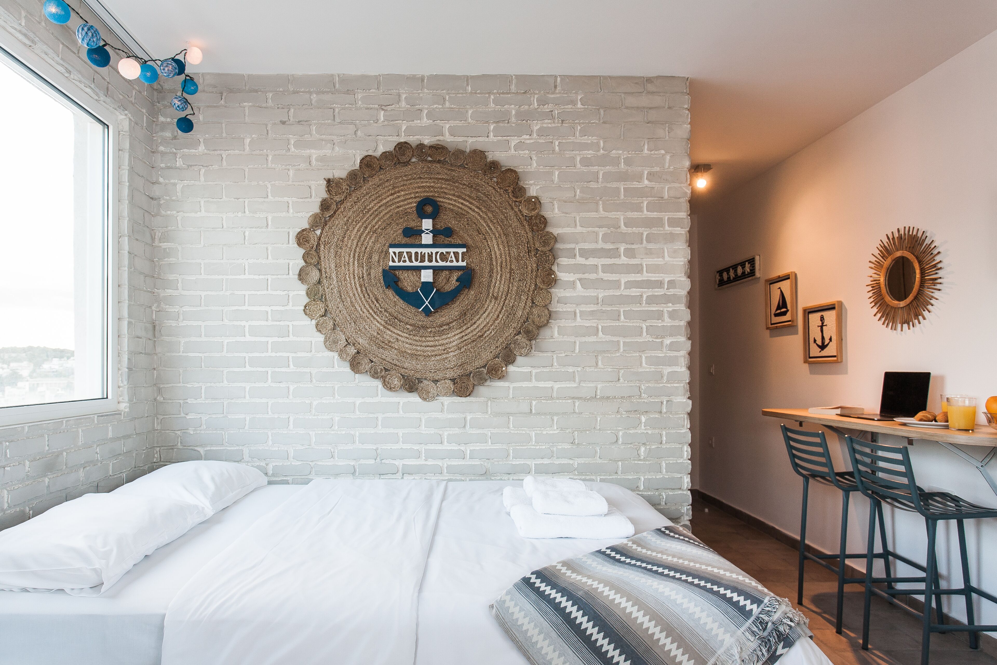Una habitación de hotel con pared de ladrillo blanco, un tapiz circular tejido con diseño de ancla, una cama con ropa de cama blanca, una manta de cuadros y una barra de desayuno con sillas.