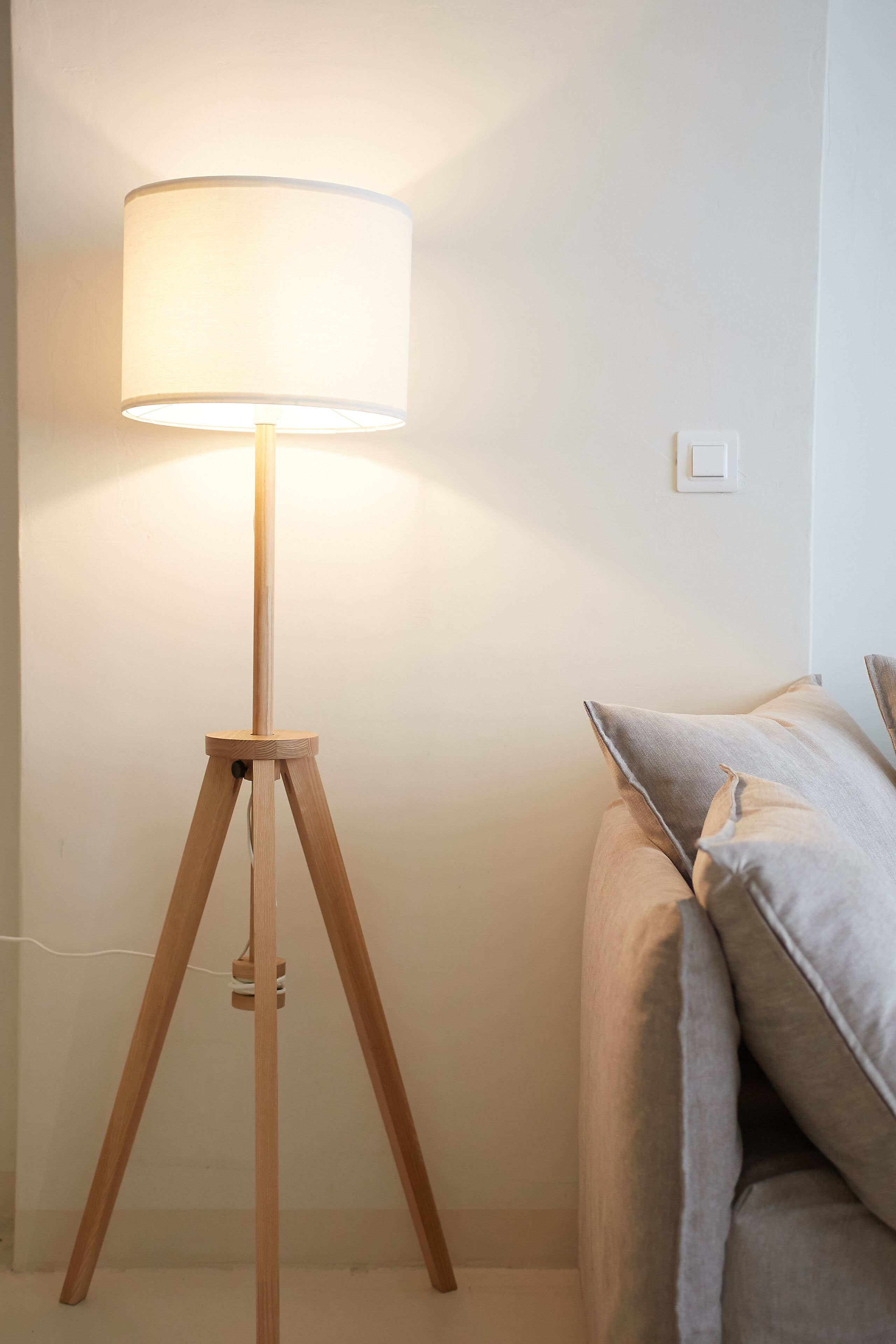 A floor lamp with a white shade on a tripod stand, positioned next to a bed with grey bedding.