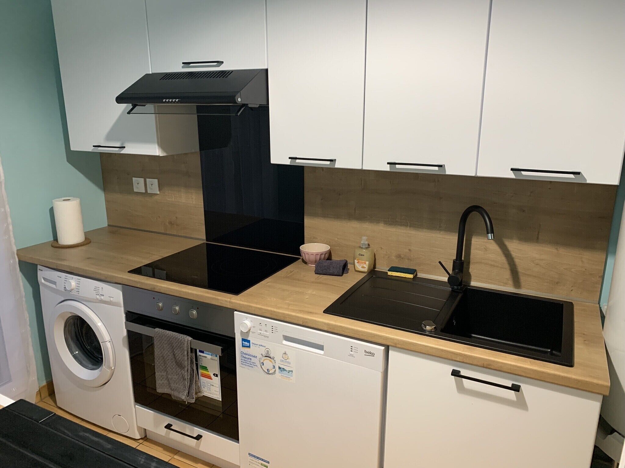 Ett kompakt kök med tvättmaskin, diskmaskin och en svart köksfläkt.