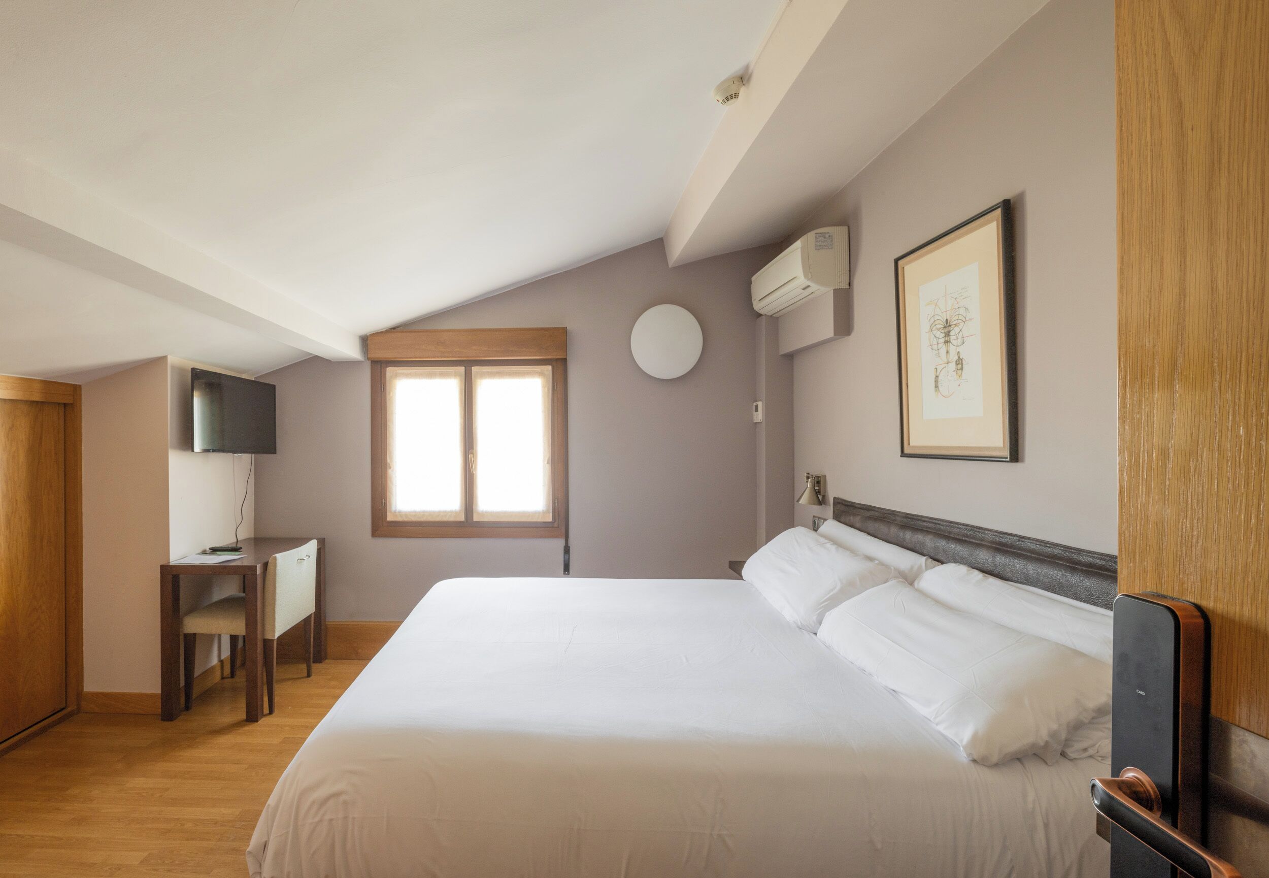 A bedroom with a large bed, a desk, a chair, a window, and a wall-mounted air conditioner.