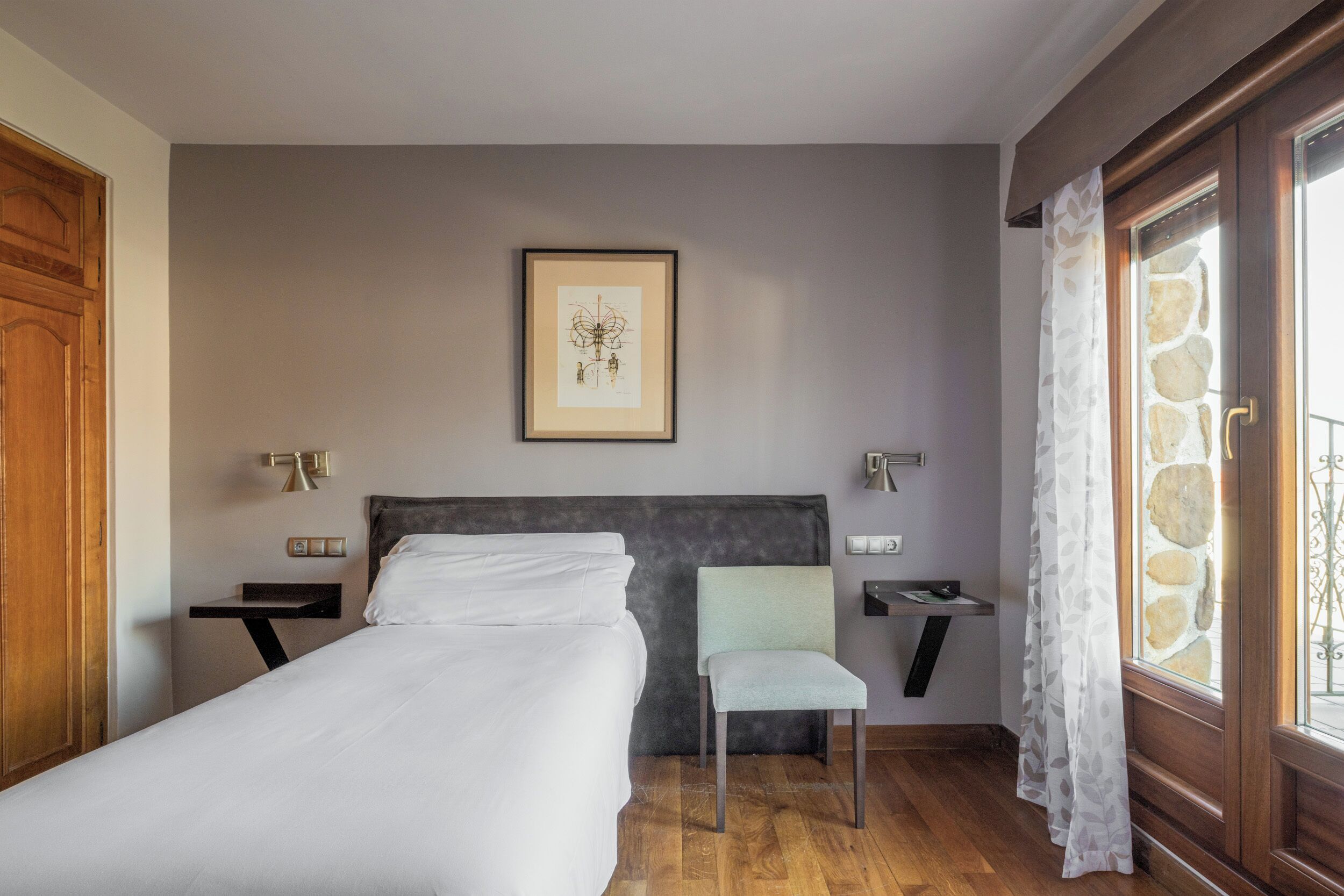 Una camera da letto ordinata con un letto, una sedia, comodini, un quadro incorniciato e una finestra con tenda.