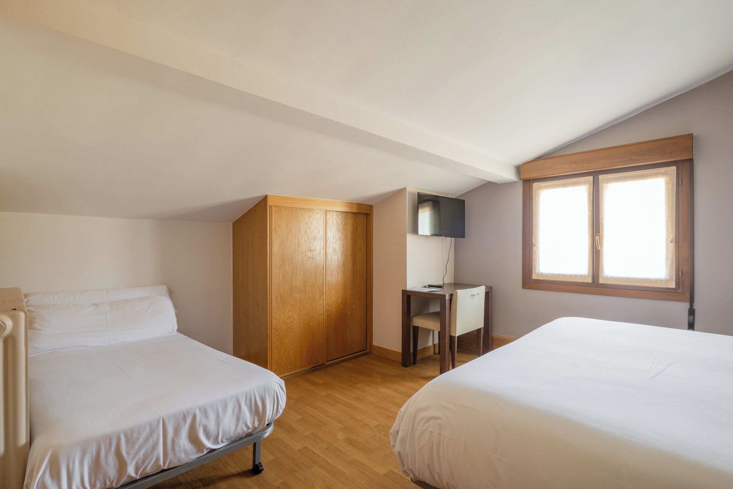 Un dormitorio con dos camas, un armario de madera, un escritorio con silla y un televisor fijado a la pared.